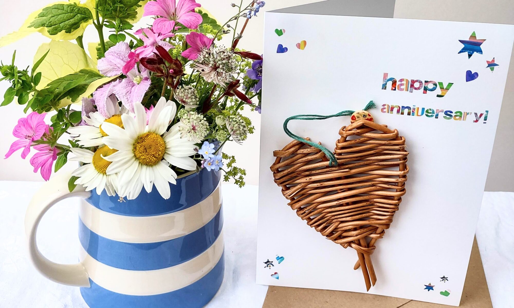 woven willow heart on a white card with rainbow coloured lettering, next to a brown envelope and a jug of flowers