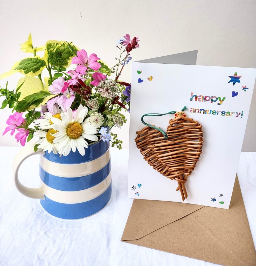 woven willow heart on a white card with rainbow coloured lettering, next to a brown envelope and a jug of flowers